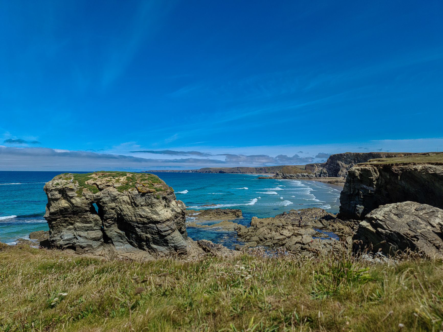 Circular coast path walk climbing a green Cornish headland above a sandy cove