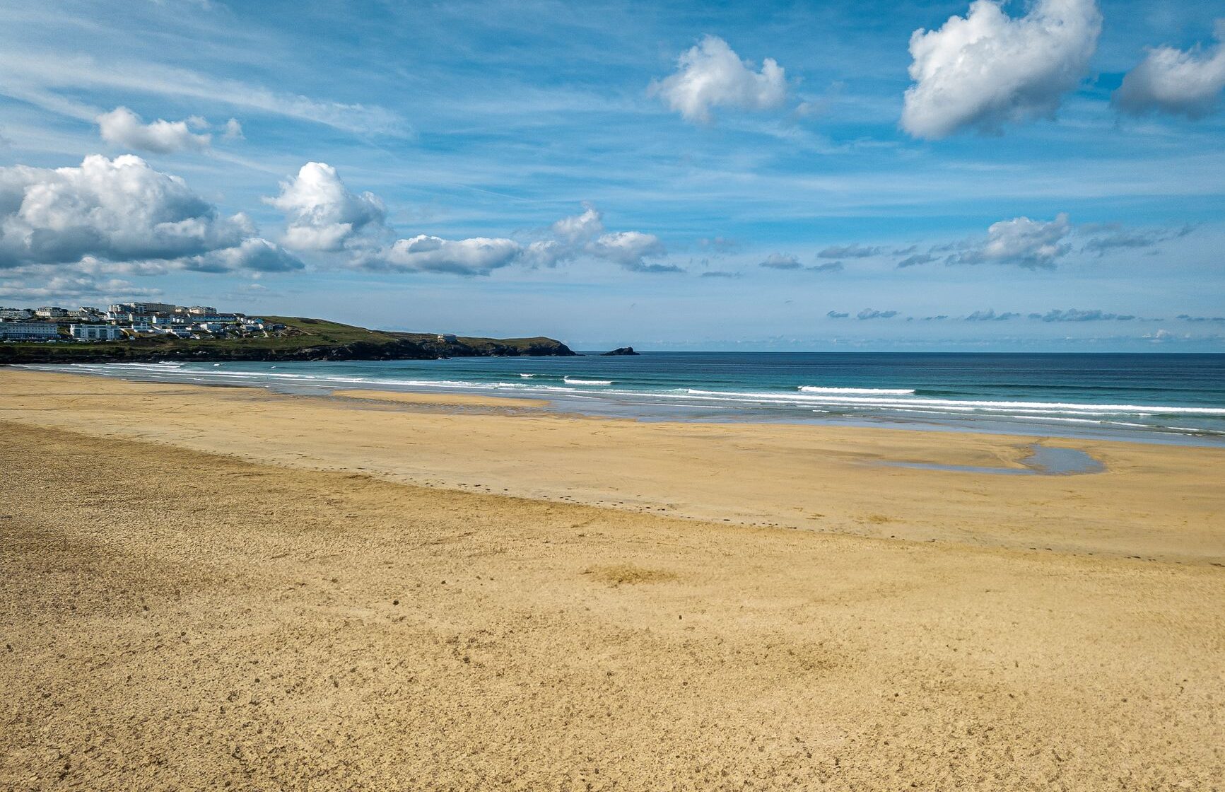 A wide, near-empty stretch of golden sand at low tide in Newquay, with a headland and small offshore rock beyond