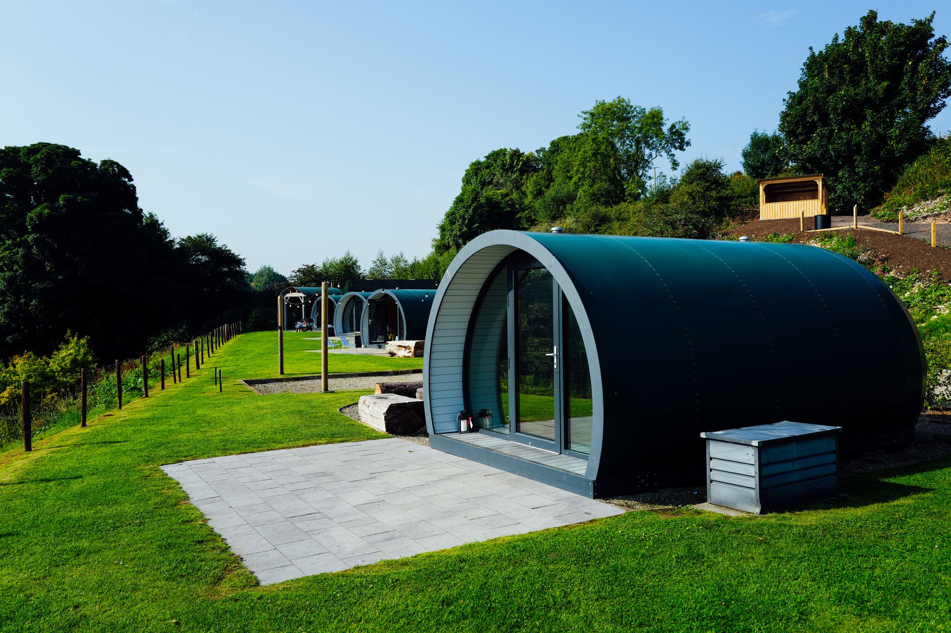 A row of wooden glamping pods on a grassy field with trees behind
