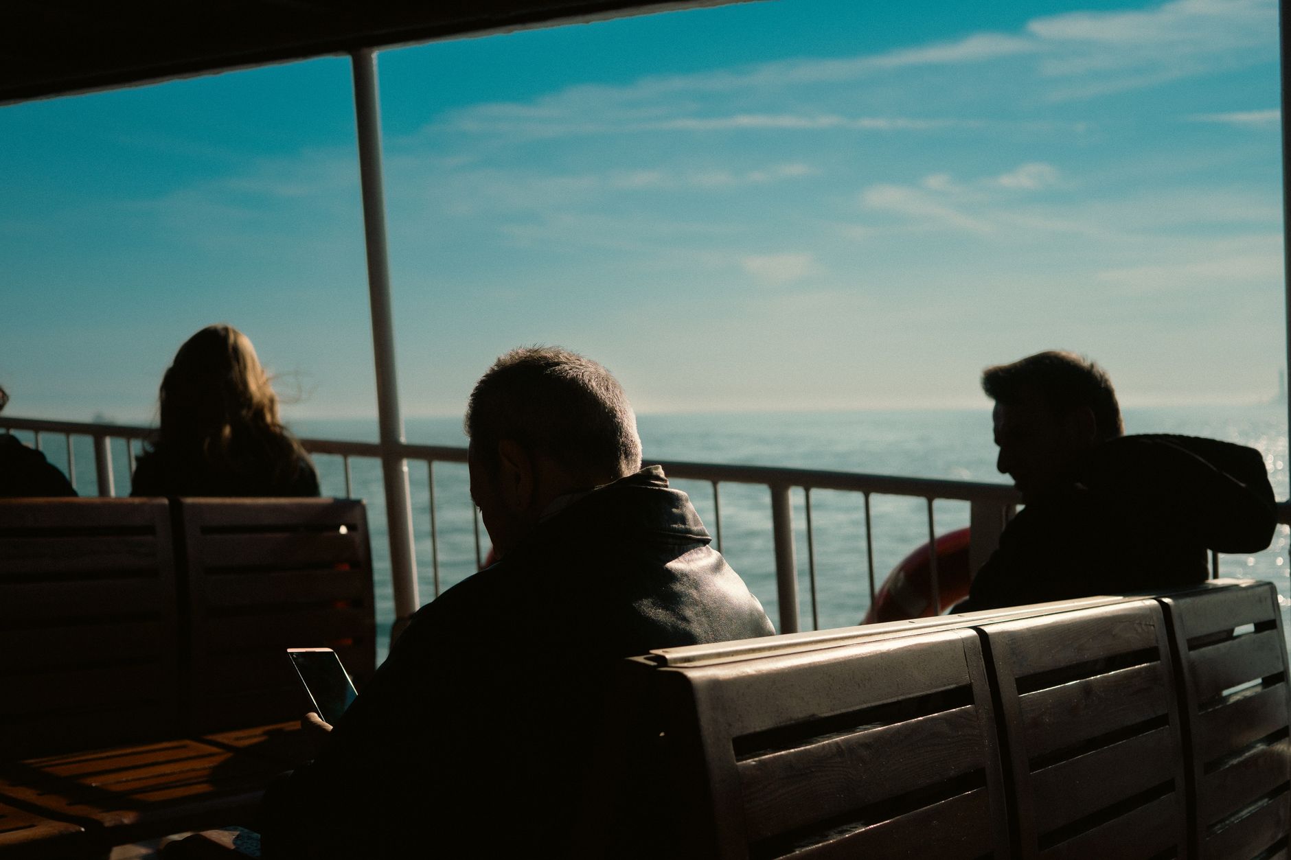 Passengers sitting on benches on a ferry's covered outside deck, looking out to sea