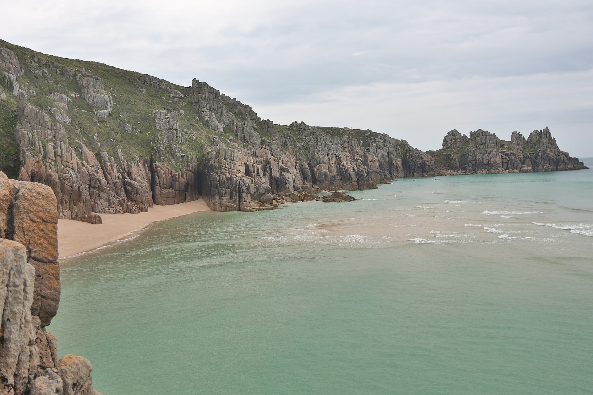 Pale sand at Pedn Vounder below granite cliffs, with the Treryn Dinas headland beyond