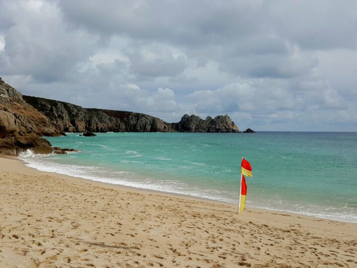 Porthcurno beach with a red and yellow lifeguard flag, turquoise sea and the Logan Rock headland beyond